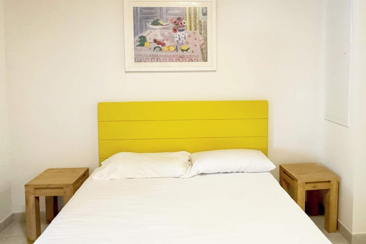 A bedroom with a yellow headboard, white bedding, two wooden nightstands, and a framed picture on the wall.