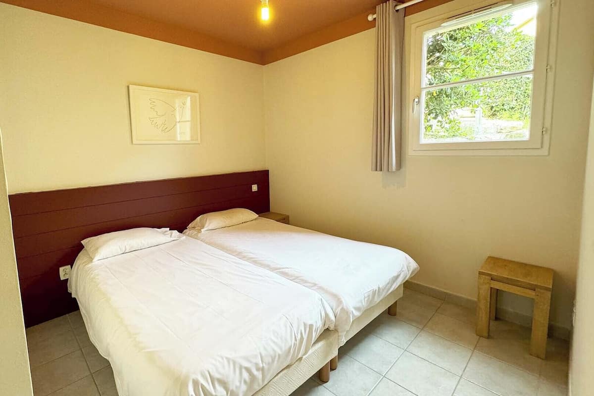 A bedroom with a bed, a nightstand, a small table, a window with curtains, and a wall-mounted picture.