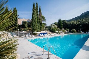 A swimming pool with lounge chairs and umbrellas, surrounded by trees and a building with arches. - Village Club du Soleil - Le Reverdi (Plan-de-la-Tour)