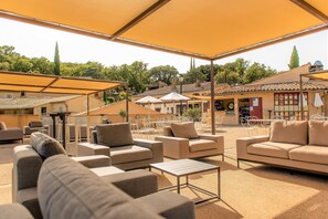 An outdoor seating area with multiple sofas and tables, shaded by large umbrellas, and a view of buildings and trees in the background. - Village Club du Soleil - Le Reverdi (Plan-de-la-Tour)