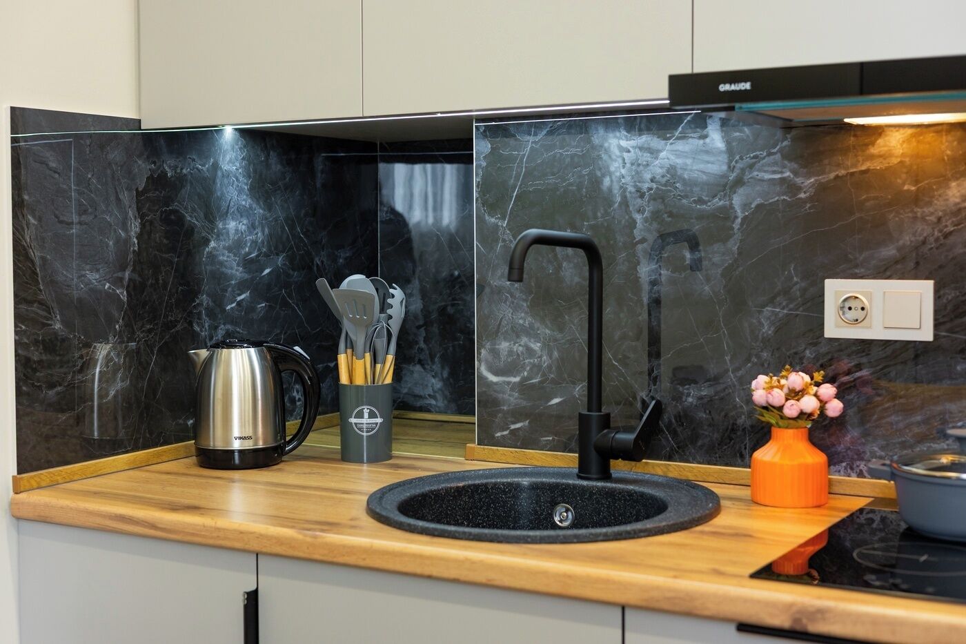 Una cocina moderna con un lavamanos negro, hervidor de acero inoxidable y un florero con flores.