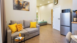 Un salon avec un canapé, un coin repas avec une table, une cuisine avec un réfrigérateur et un couloir menant à une chambre.