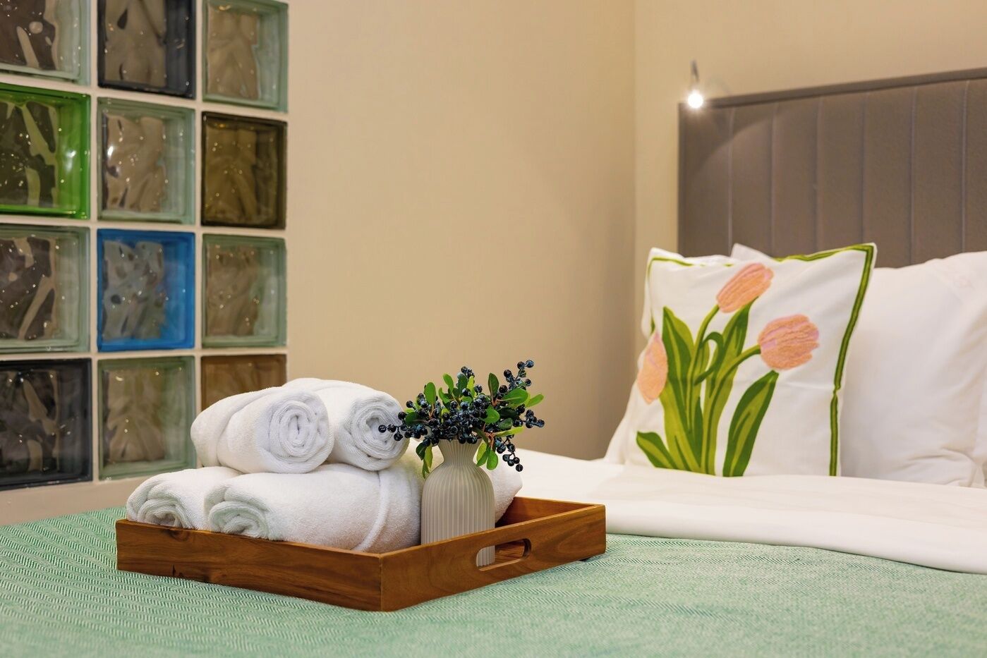 Una cama bien hecha con ropa blanca y una almohada con diseño de tulipán, una bandeja de madera con toallas dobladas y un florero con follaje, y una pared con obras de arte enmarcadas.