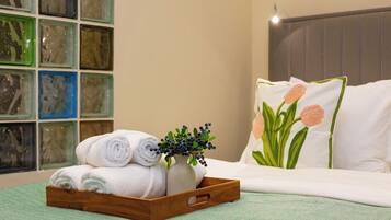Una cama bien hecha con ropa blanca y una almohada con diseño de tulipán, una bandeja de madera con toallas dobladas y un florero con follaje, y una pared con obras de arte enmarcadas.