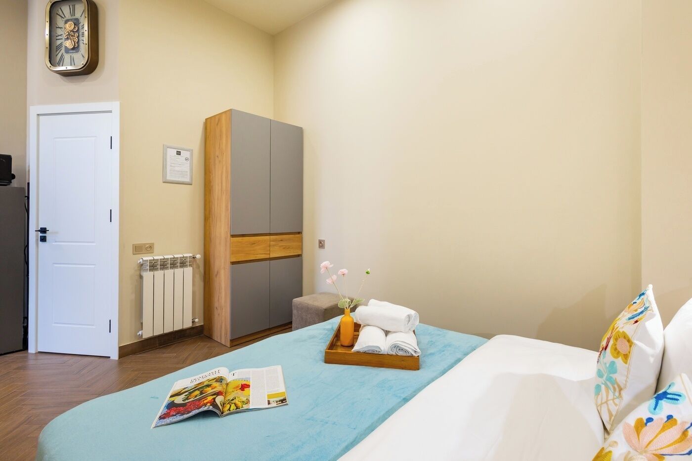 A hotel room with a bed, a wooden wardrobe, a radiator, a clock, and a small table with towels and a magazine.