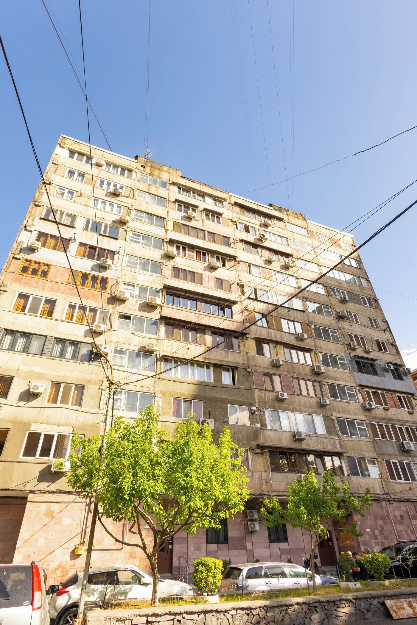 Un edificio a più piani con numerose finestre e condizionatori, circondato da alberi e auto parcheggiate.