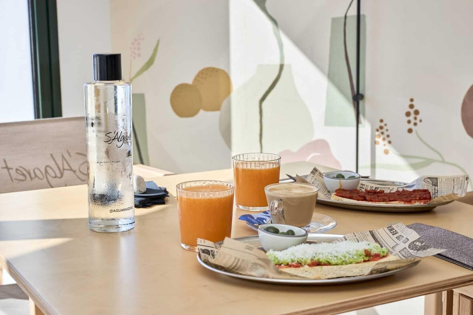 A breakfast setting with a bottle of water, two glasses of orange juice, a cup of coffee, and a plate with a sandwich and a bowl of green olives.