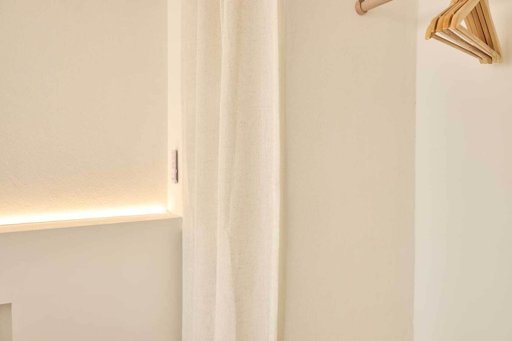 Une pièce avec un mur blanc, un rideau blanc et une étagère en bois sur laquelle se trouve un petit objet.