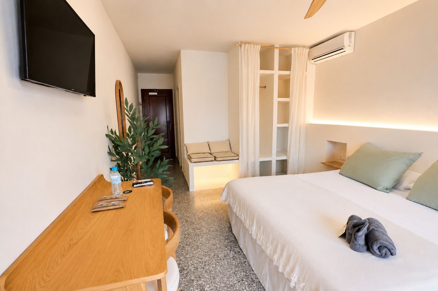 A hotel room with a bed, a desk, a TV, a plant, and an air conditioning unit.