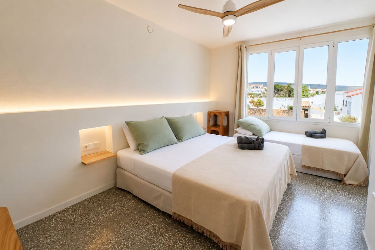 A bedroom with a bed, a bench, a ceiling fan, and a window with a view.