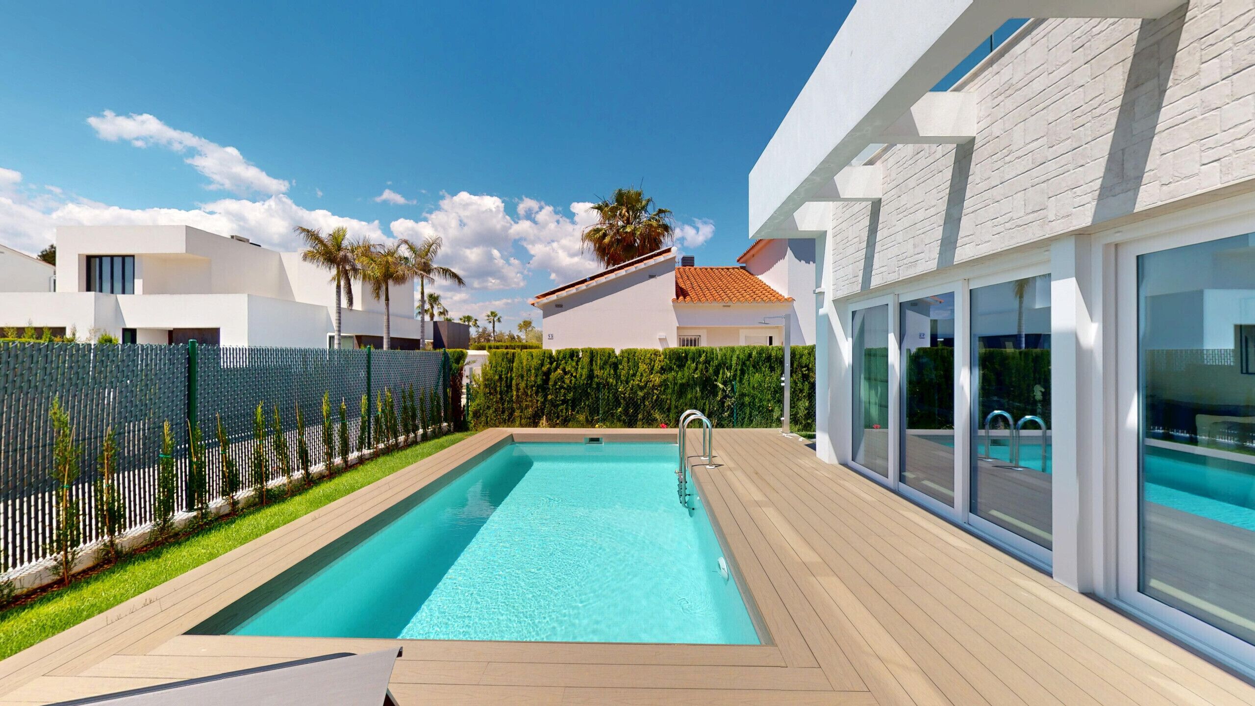 A modern swimming pool with a wooden deck, surrounded by a white building with large windows and a green fence.