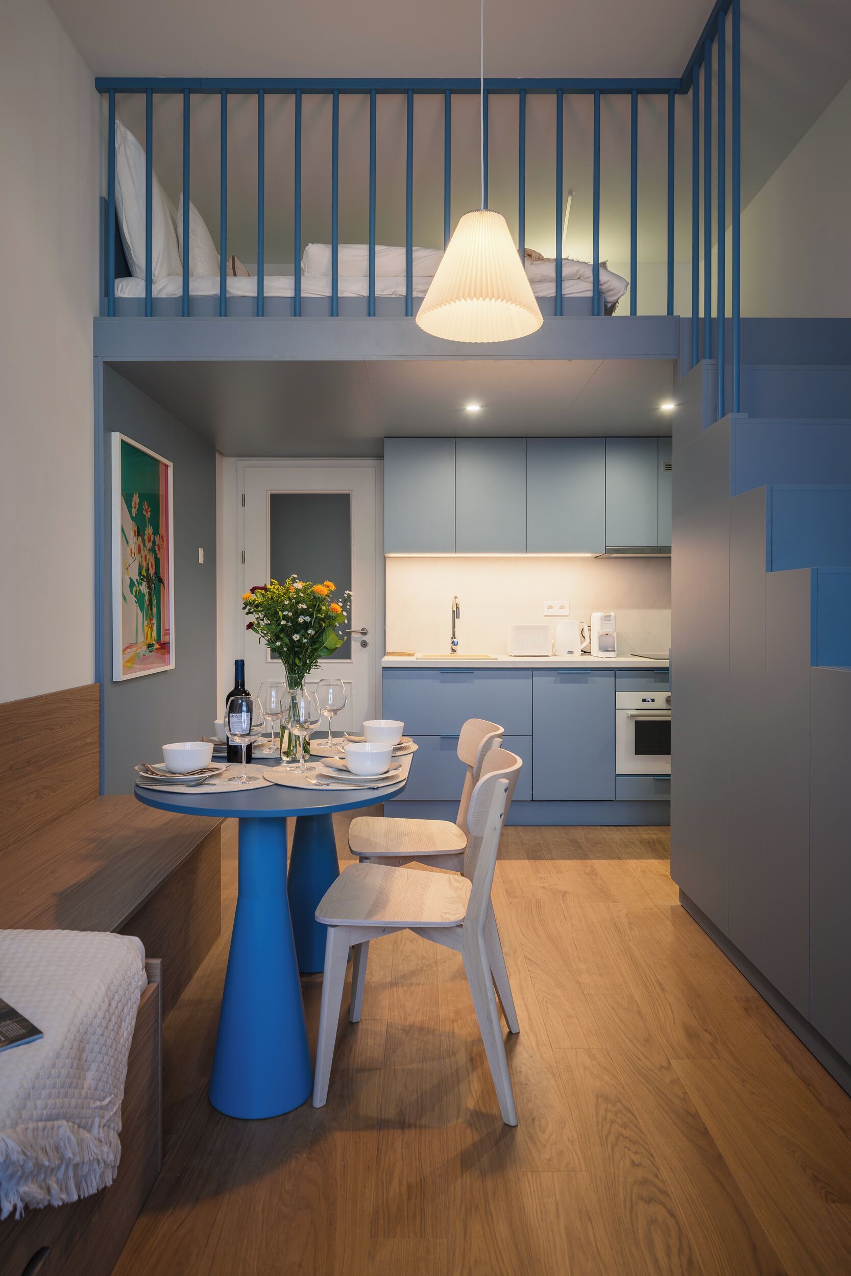 Une cuisine moderne avec des meubles bleus, un coin repas doté d’une table bleue et un escalier menant à une chambre à coucher située en mezzanine.