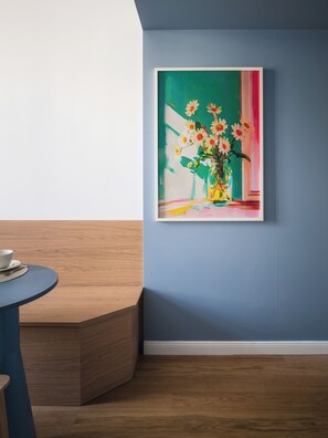 A room with a blue wall, a wooden floor, a round table with a bowl and a spoon, and a framed painting of flowers. - Designer attic studio with smart TV and WiFi (Hlavní město Praha)
