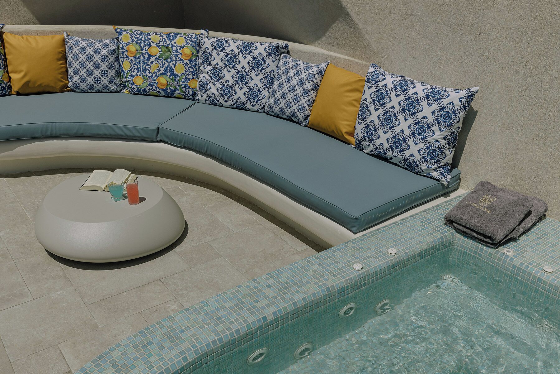 A curved outdoor seating area with a pool, a round table with a book and drink, and a towel on the poolside.
