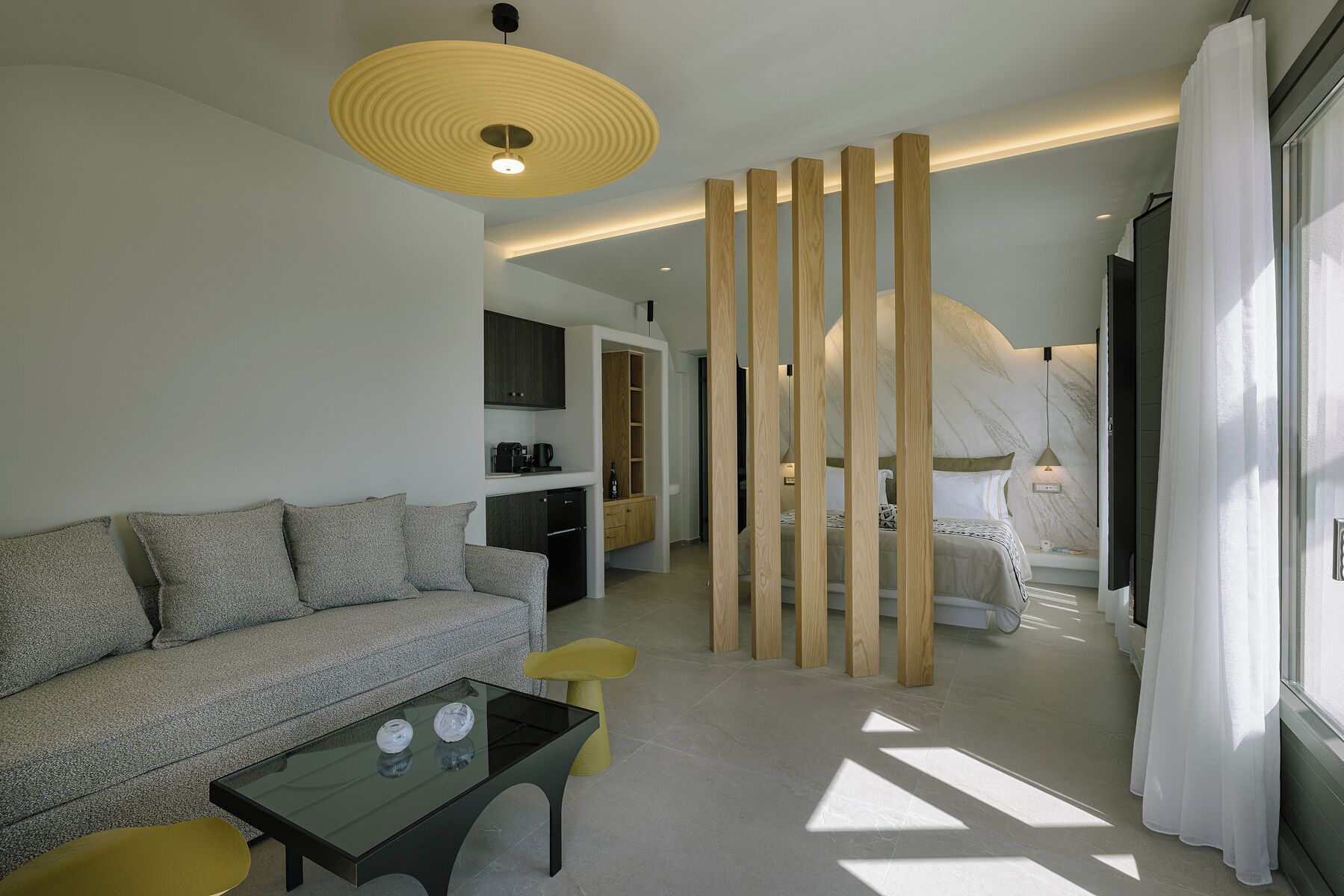 Een moderne hotelkamer met een bank, een glazen bijzettafeltje, een houten scheidingswand, een bed en uitzicht op buiten.