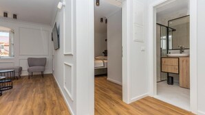 Een modern interieur met een houten vloer, een grijze fauteuil, een klein tafeltje, een bed en een badkamer met een spiegel en wastafel.