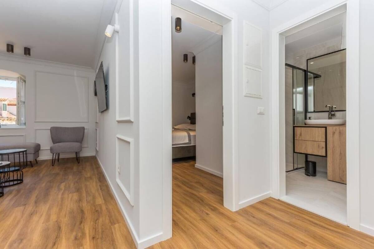 A modern interior with a wooden floor, a grey armchair, a small table, a bed, and a bathroom with a mirror and sink.