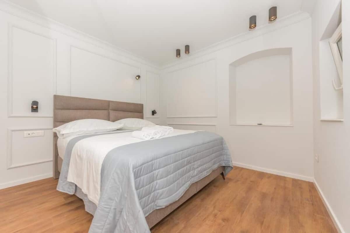 A bedroom with a bed, white walls, wooden flooring, and a ceiling light fixture.