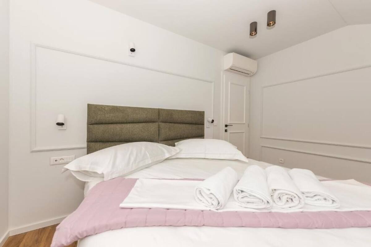 A neatly made bed with white linens and pink accents, a headboard, and a wall-mounted air conditioner.