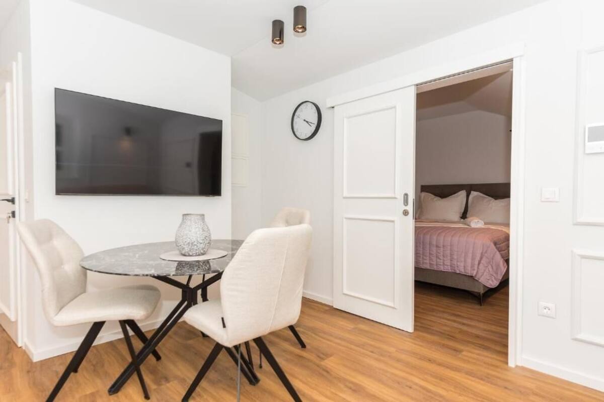 A room with a round table, two chairs, a TV, a clock, and a bed with a pink blanket.