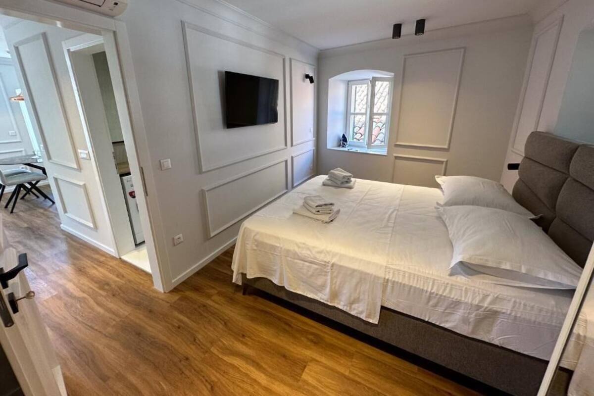 A bedroom with a bed, a television mounted on the wall, a window, and a door leading to another room.