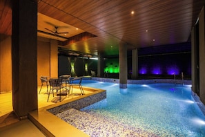 An indoor swimming pool with a tiled floor, a seating area with chairs, and a ceiling fan.