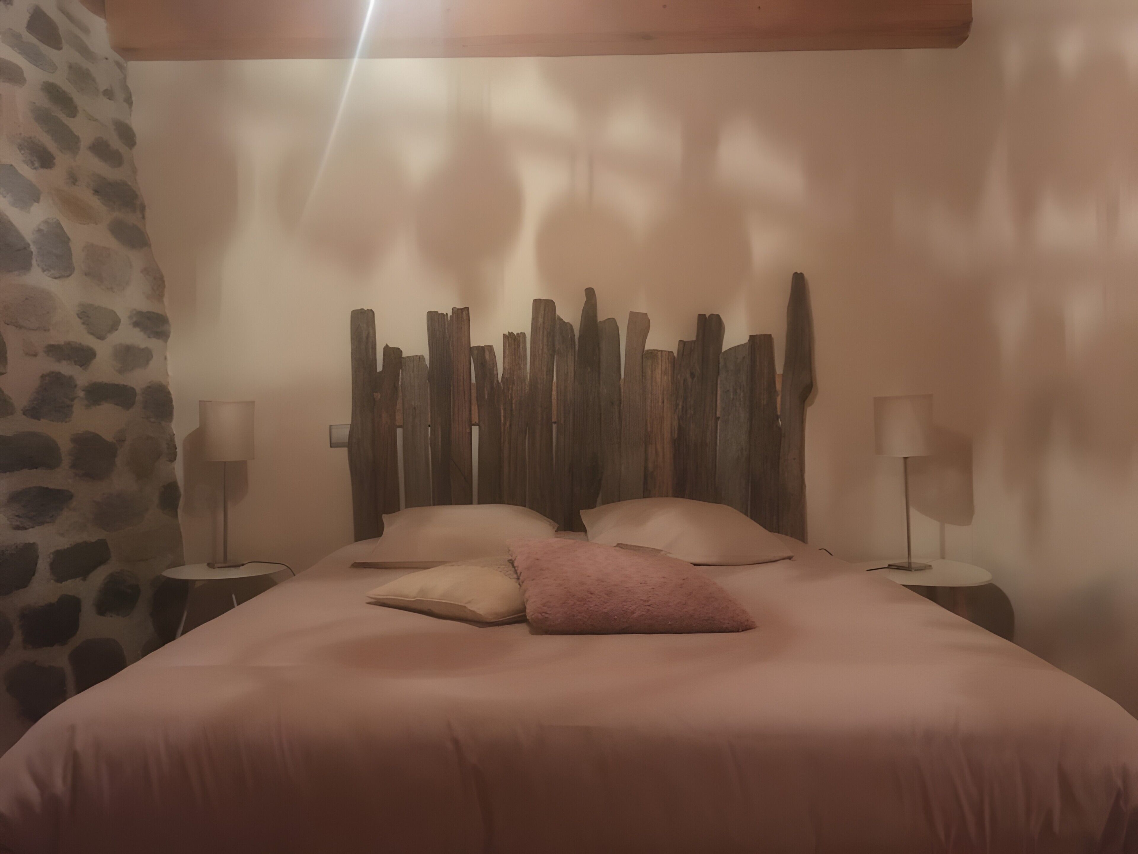 Ett sovrum med en vägg av sten, en sänggavel i trä, en säng med rosa sängkläder och två sänglampor.
