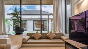 Un salón moderno con un televisor grande, un sofá con cojines, una planta en maceta y vistas a una casa a través de amplias ventanas.