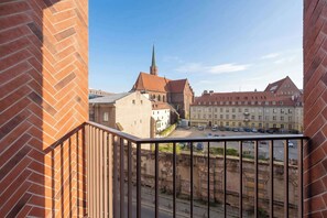 Blick vom Balkon auf ein historisches Gebäude mit einem markanten Turm, eine gepflasterte Straße und abgestellte Autos.