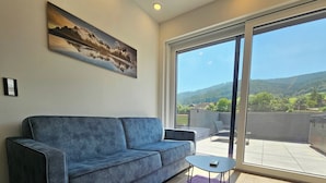 A modern living room with a blue sofa, a small table, and a large sliding glass door leading to a balcony with a scenic mountain view. - Lorenzer Penthouse (Scheifling)