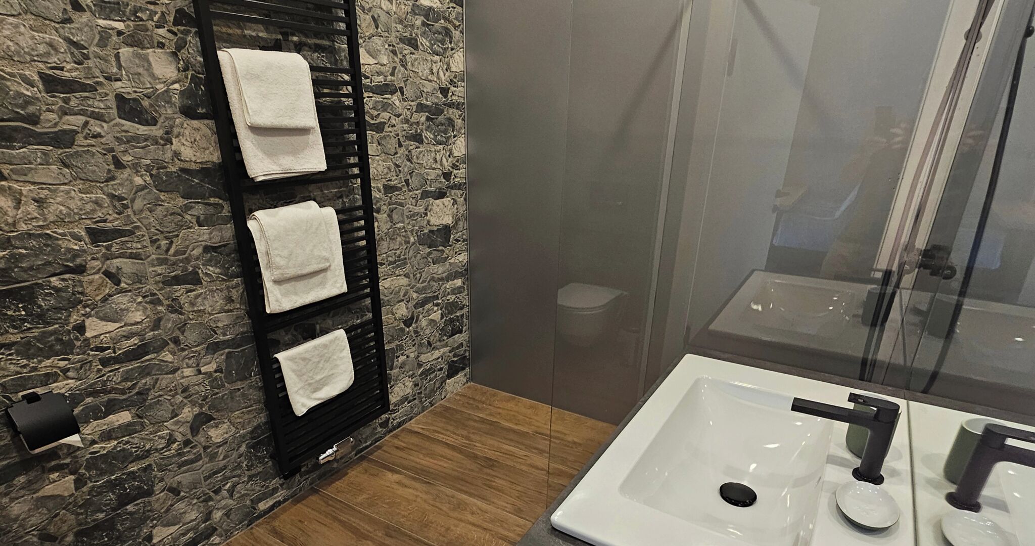 A bathroom with a stone wall, a black towel rack with three towels, a wooden floor, a white sink with a black faucet, and a glass shower enclosure.