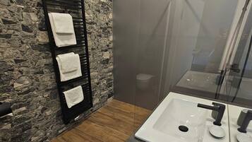 A bathroom with a stone wall, a black towel rack with three towels, a wooden floor, a white sink with a black faucet, and a glass shower enclosure.