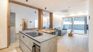 A modern kitchen with a central island, a dining area with a wooden table, and a living space with a sofa and a large window. - Lorenzer Penthouse (Scheifling)