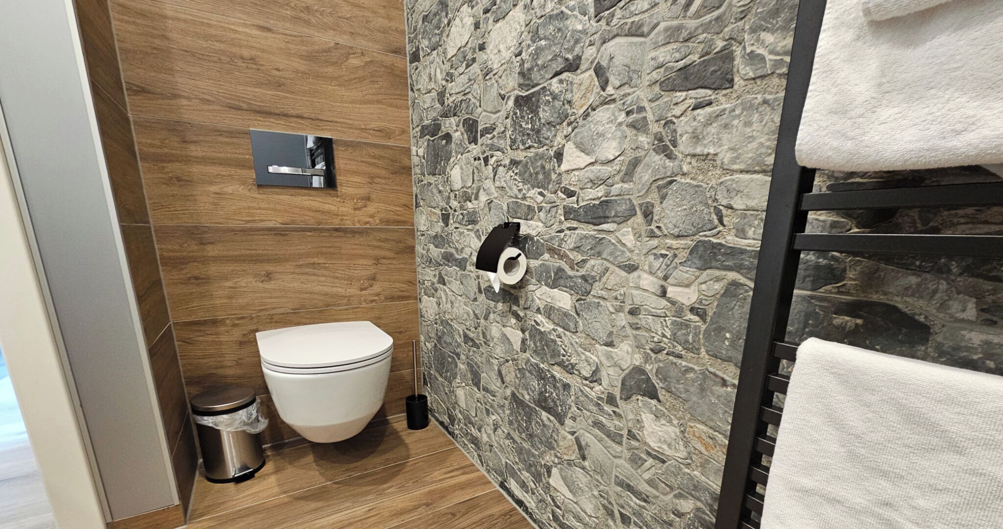 A bathroom with a toilet, a trash bin, a towel rack, and a stone-patterned wall.