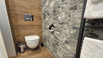 A bathroom with a toilet, a trash bin, a towel rack, and a stone-patterned wall.