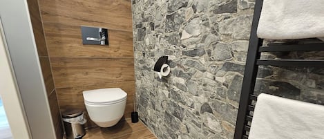 A bathroom with a toilet, a trash bin, a towel rack, and a stone-patterned wall.