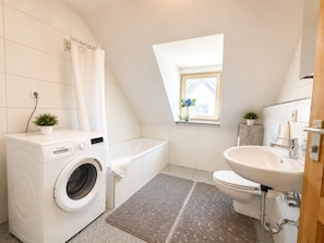 Un baño moderno y funcional con lavadora Bosch, bañera, inodoro y una ventana pequeña con vistas.