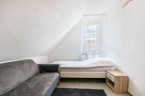 Una habitación pequeña y moderna con cama, sofá, mesita de noche y ventana con cortinas translúcidas.