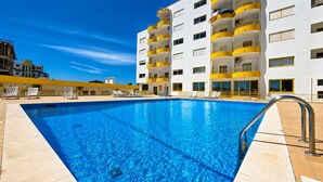 A swimming pool with a clear blue water, surrounded by a white building with yellow balconies, and a few lounge chairs. - Mar & Sol Penthouse Portimão with Sea View, Wi-Fi, and Air Conditioning (Portimão)
