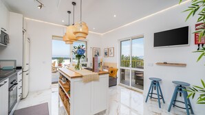 A modern kitchen with a central island, a dining area with blue stools, a flat-screen TV, and a large window offering a view. - Mar & Sol Penthouse Portimão with Sea View, Wi-Fi, and Air Conditioning (Portimão)