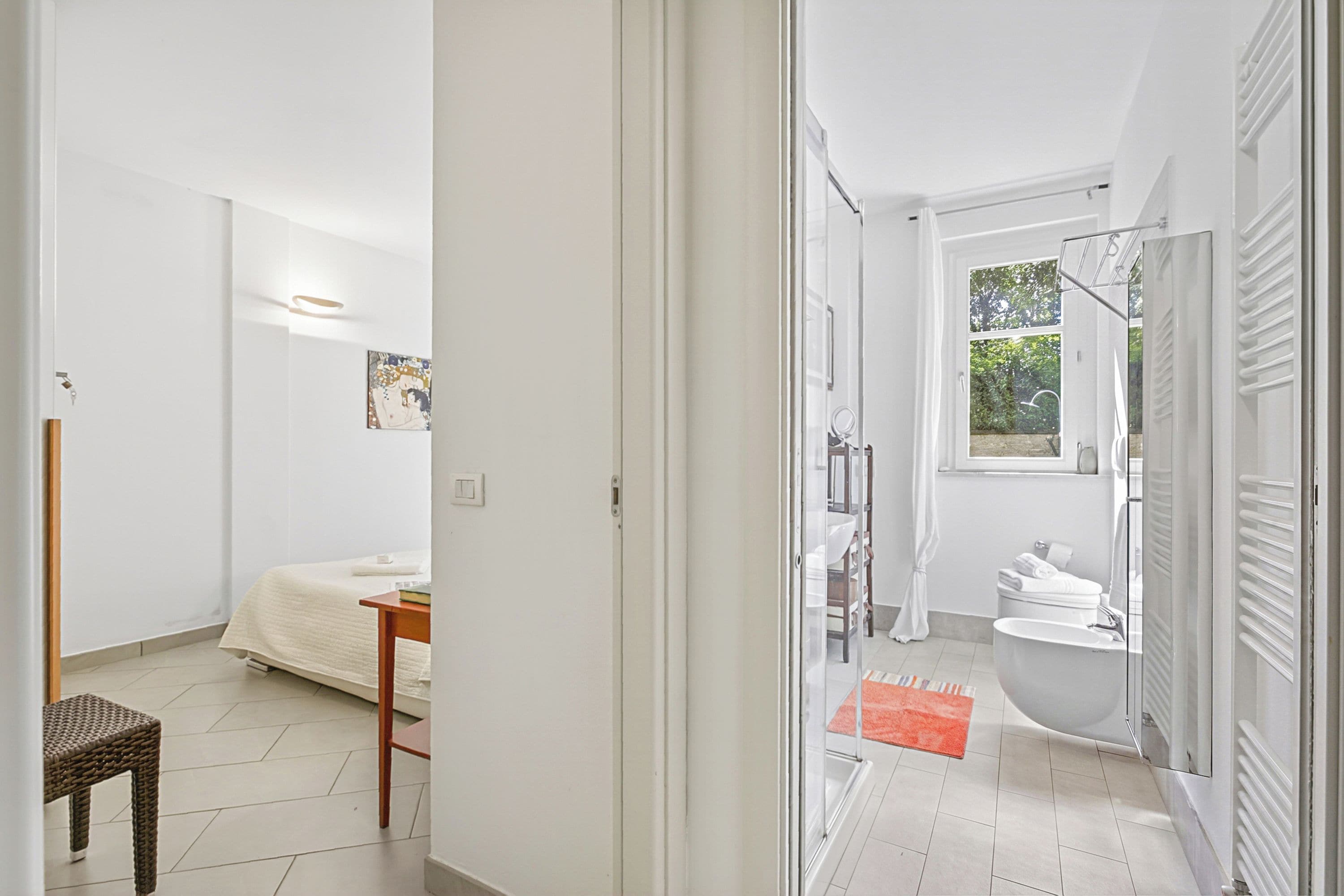 Une salle de bain moderne avec une baignoire, des toilettes et un espace douche, un petit coin salon avec une chaise et une table, et un lit dans la pièce adjacente.