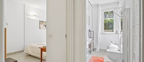 A modern bathroom with a bathtub, toilet, and shower area, a small seating area with a chair and table, and a bed in the adjacent room.