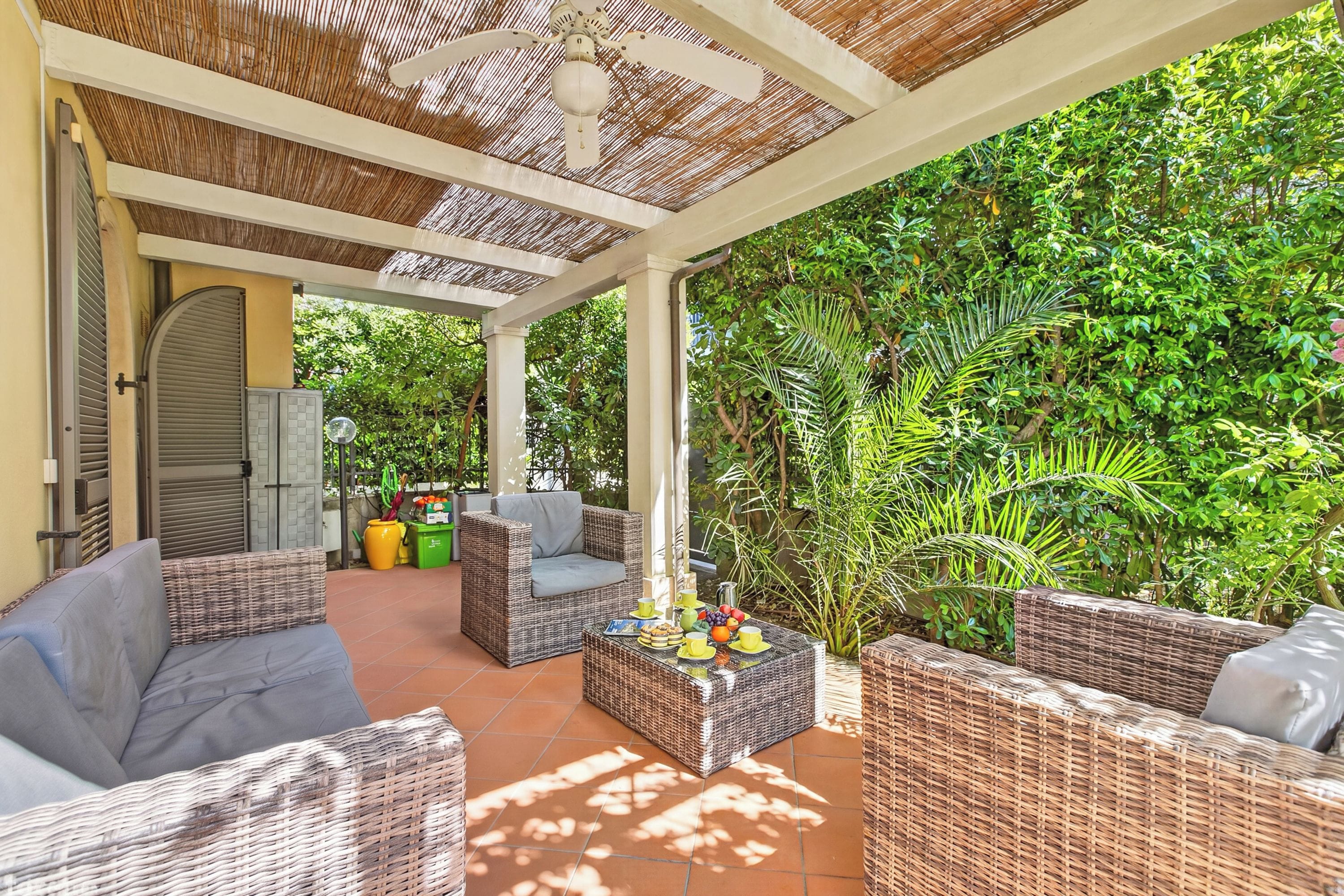 A covered outdoor seating area with wicker furniture and a table set with fruits and cups.