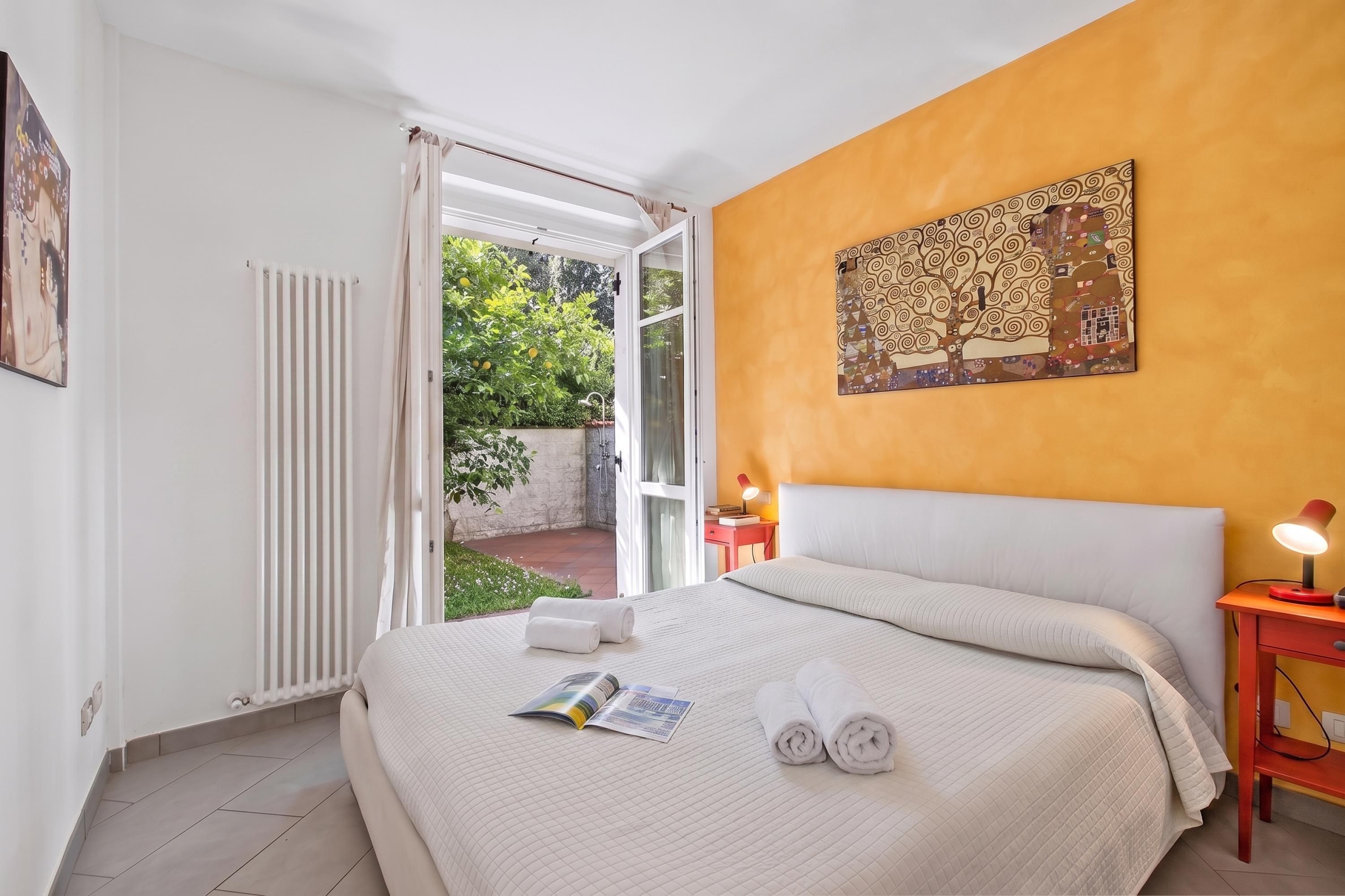 A hotel room with a bed, a reading area, a nightstand, a window with curtains, and a painting on the wall.
