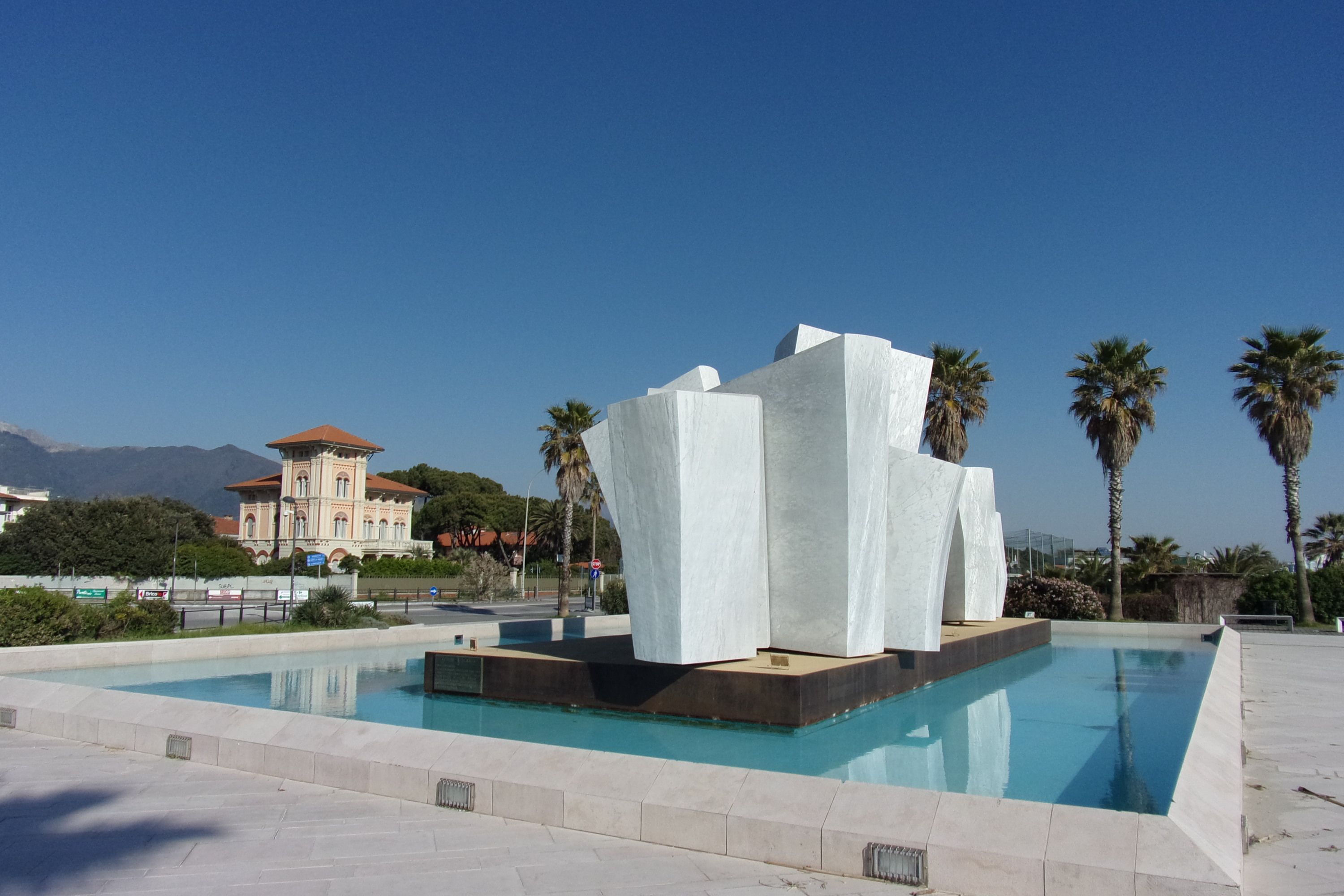 A modern pool area with a unique white sculpture centerpiece, surrounded by palm trees and a clear blue sky.