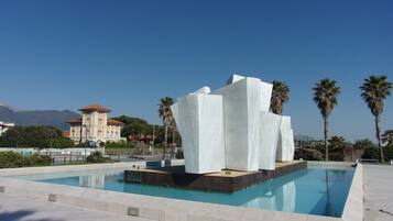 A modern pool area with a unique white sculpture centerpiece, surrounded by palm trees and a clear blue sky.