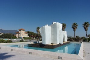 A modern pool area with a unique white sculpture centerpiece, surrounded by palm trees and a clear blue sky. - Maison Clelie With Garden - 200 M From Sea (Massa)