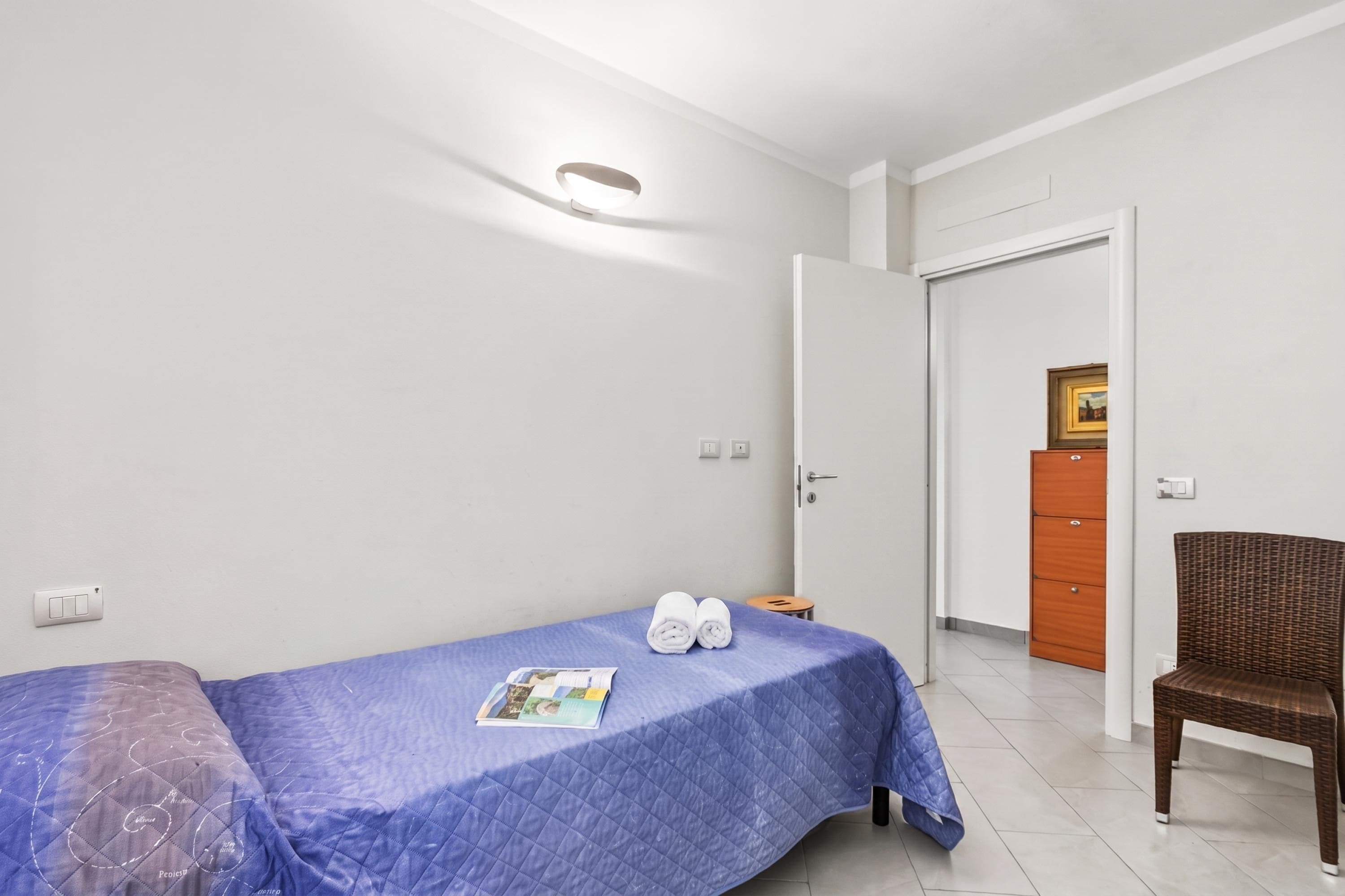 A single bed with a blue bedspread, a pair of rolled towels, and a magazine on the bed. There is a wooden chair and a wardrobe in the room.
