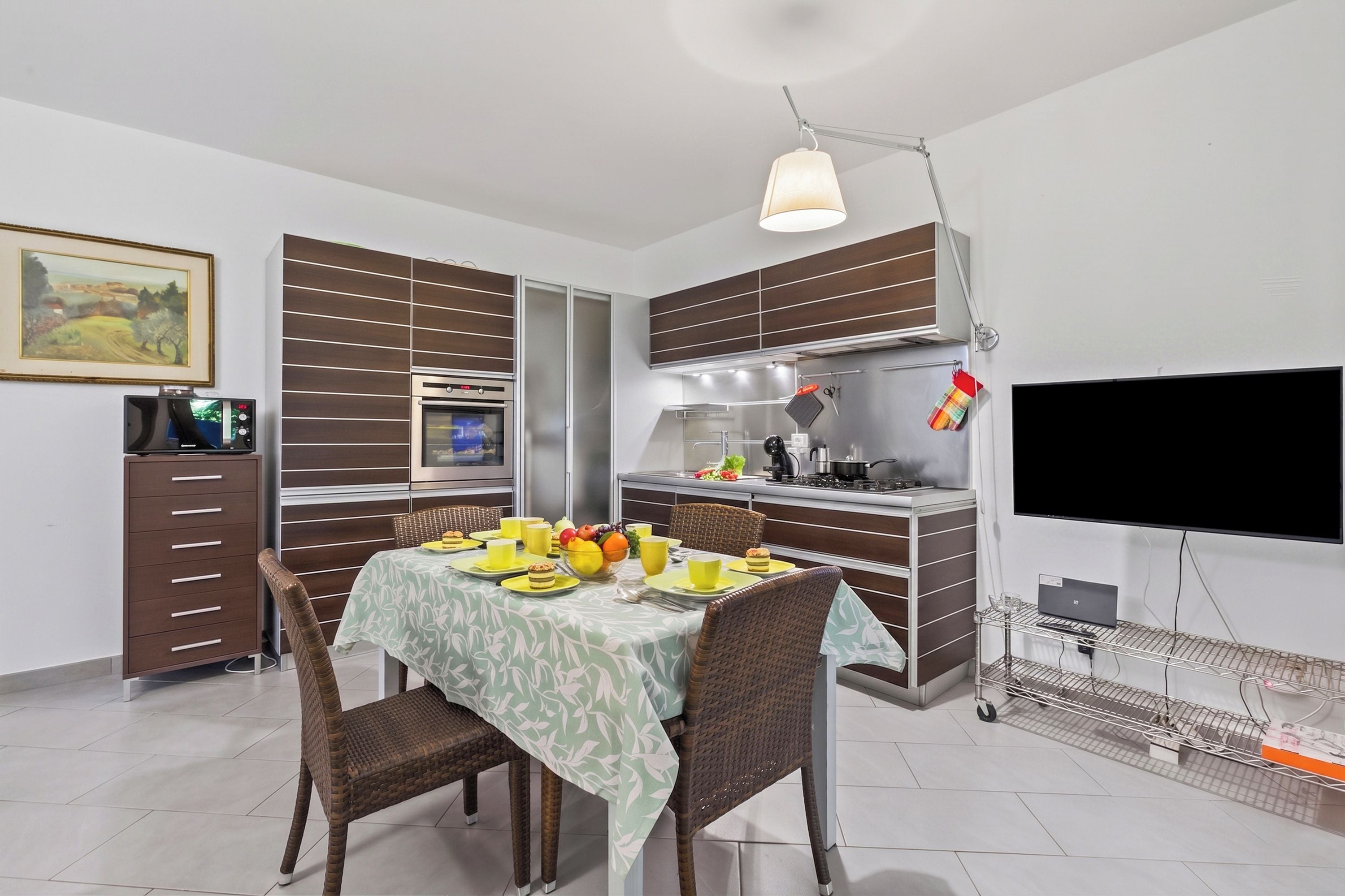 A modern kitchen with a dining area, a flat-screen TV, and a laptop on a cart.