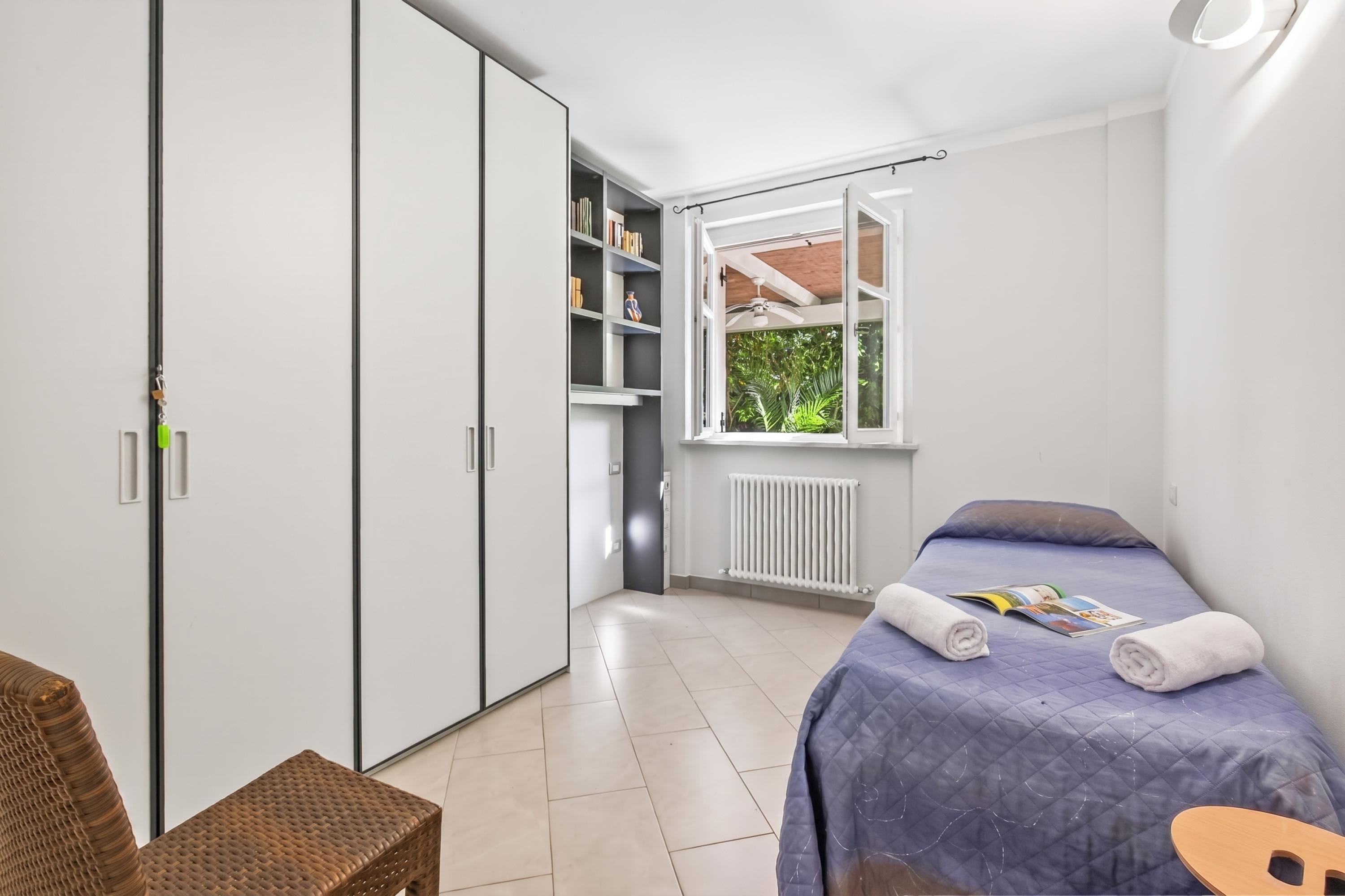 A bedroom with a bed, a window, a radiator, a bookshelf, and a wardrobe.
